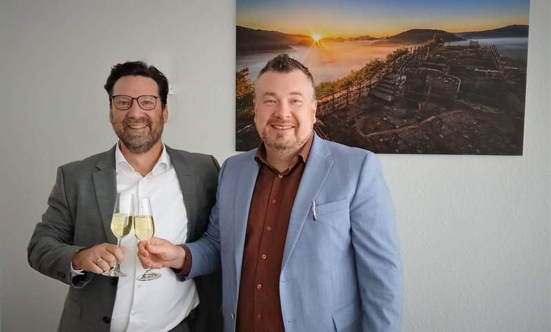 Holger Zwick (links) und Patrick Weißler bekräftigen mit (alkoholfreiem) Sekt den Grundsatzbeschluss für ein interkommunales Medizinisches Versorgungszentrum. (Wandbild zeigt Ruine Falkenburg oberhalb Wilgartswiesen, VG Hauenstein). Foto. W. G. Stähle.