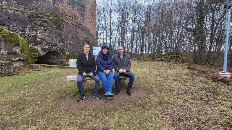 Auf der neuen Sitzbank in Ramberg haben Christian Burkhart, Bürgermeister der VG Annweiler, Ortsbürgermeisterin Marina Fess und Landrat Dietmar Seefeldt (von links nach rechts) probeweise Platz genommen. Foto. Ortsgemeinde Ramberg. Frei.
