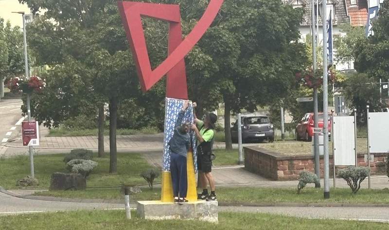 Gegen 13 Uhr. Eine Frau und ein Mann in Zimmermanns-Kluft verhüllen die rund 2 Meter hohe, in der Nacht auf Sonntag aufgesprühte Deutschland-Flagge nebst den Bundesadlern. An der Skulptur am Bahnhofs-Kreisel in BZA. Foto. Privat.